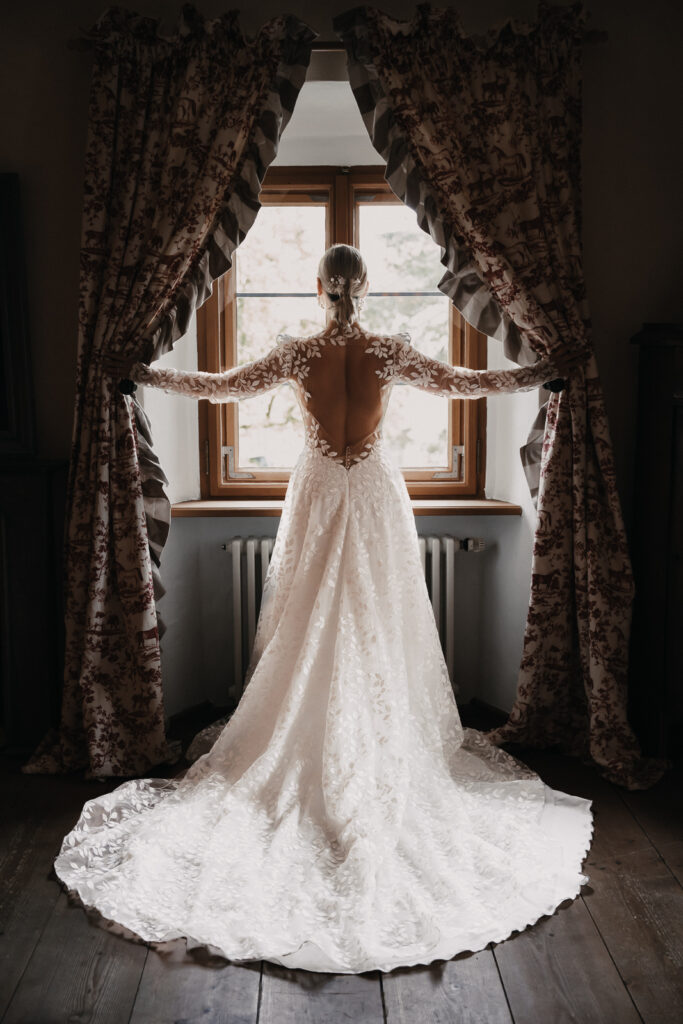 Braut steht am Fenster und man sieht ihren schönen Rücken und eine tolle, weiße Schleppe.