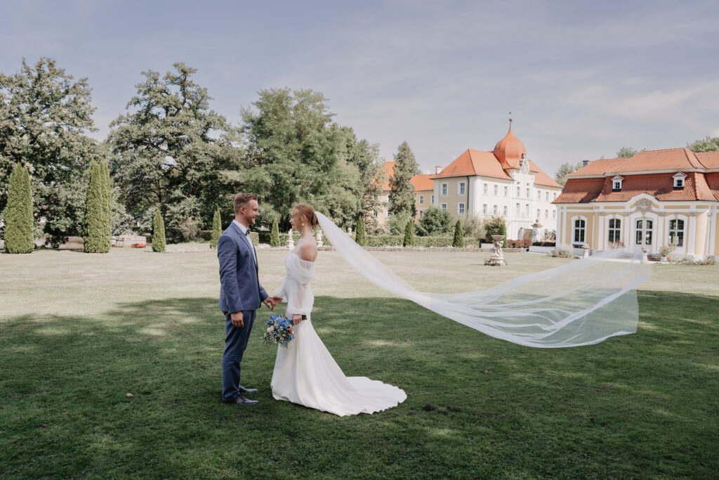 Eine Braut mit langem Schleier und ihr Bräutigam stehen Hand in Hand vor dem Schloss Thurn in Heroldsbach.